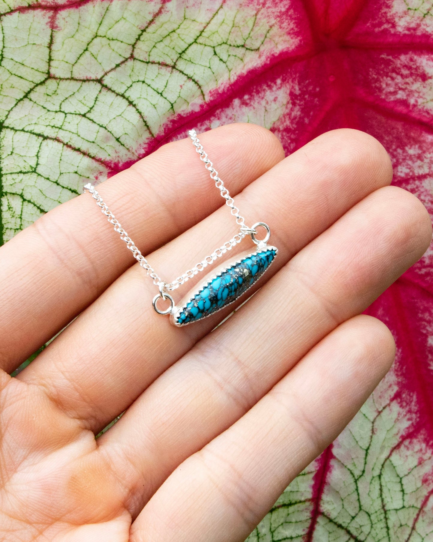 mermaid's scale horizontal marquise stone sterling silver necklace being held in front of red vains of white and green leaf