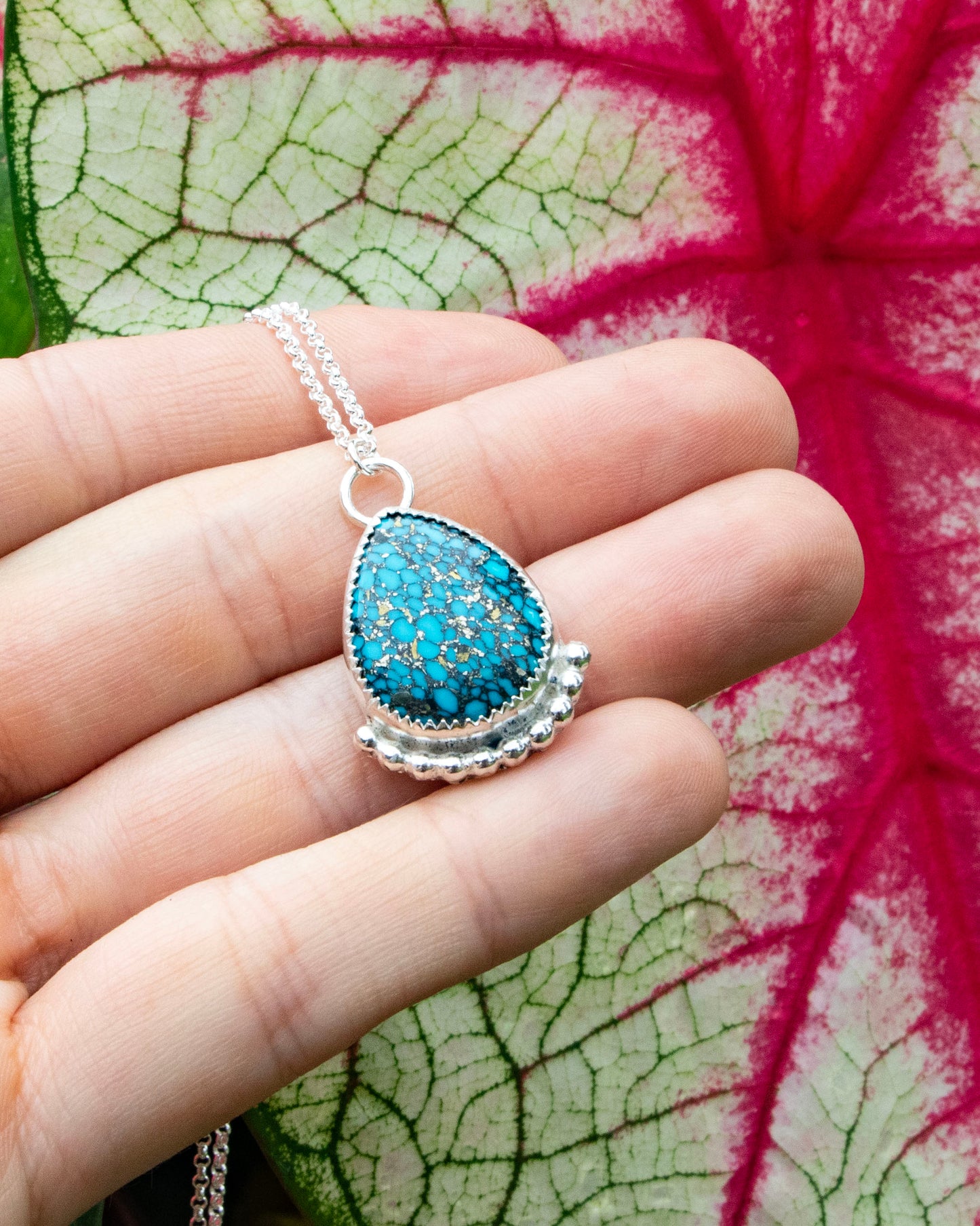 mermaid's scale teardrop stone sterling silver necklace with silver beads along bottom being held in front of red vains of white and green leaf