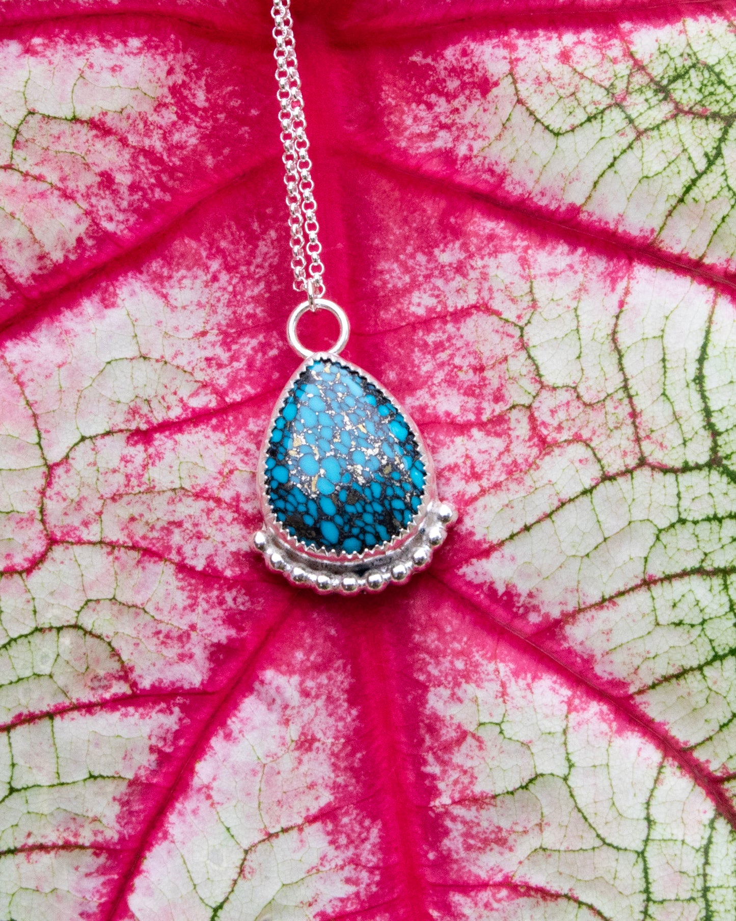 mermaid's scale teardrop stone sterling silver necklace with silver beads along bottom in front of red vains of white and green leaf