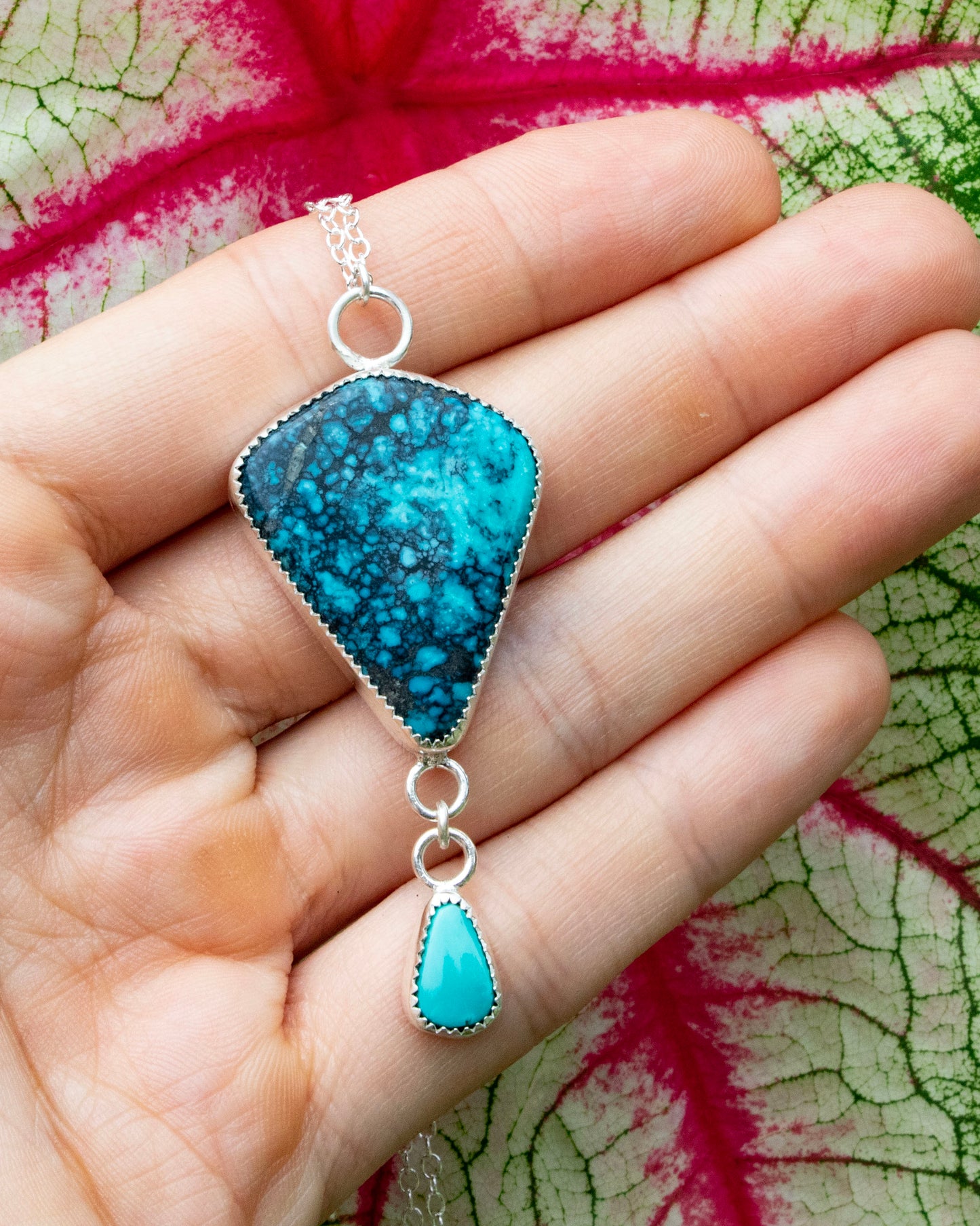 Moon River Turquoise kite sterling silver necklace with carico lake turquoise accent hung from a short jump ring chain at bottom of moon rivere stone. Necklace is being held in front of red veins of white and green leaf
