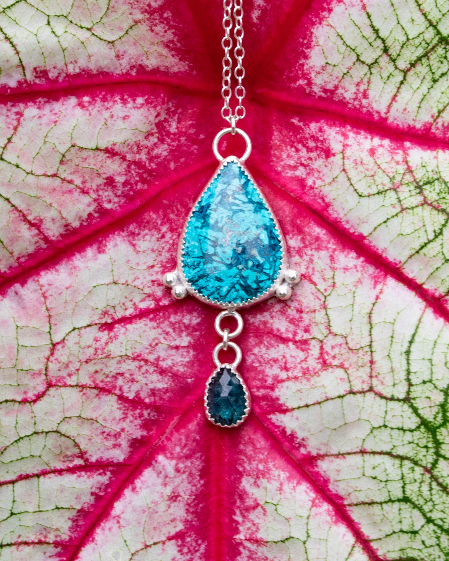 sterling silver necklace with teardrop Chrysocolla with light and dark teal blue striation pattern with two silver beads at the bottom corneres of stone. 3 jumprings connect a small teal moss kyanite hangs from the bottom. Necklace is in front of red veins on white and green leaf