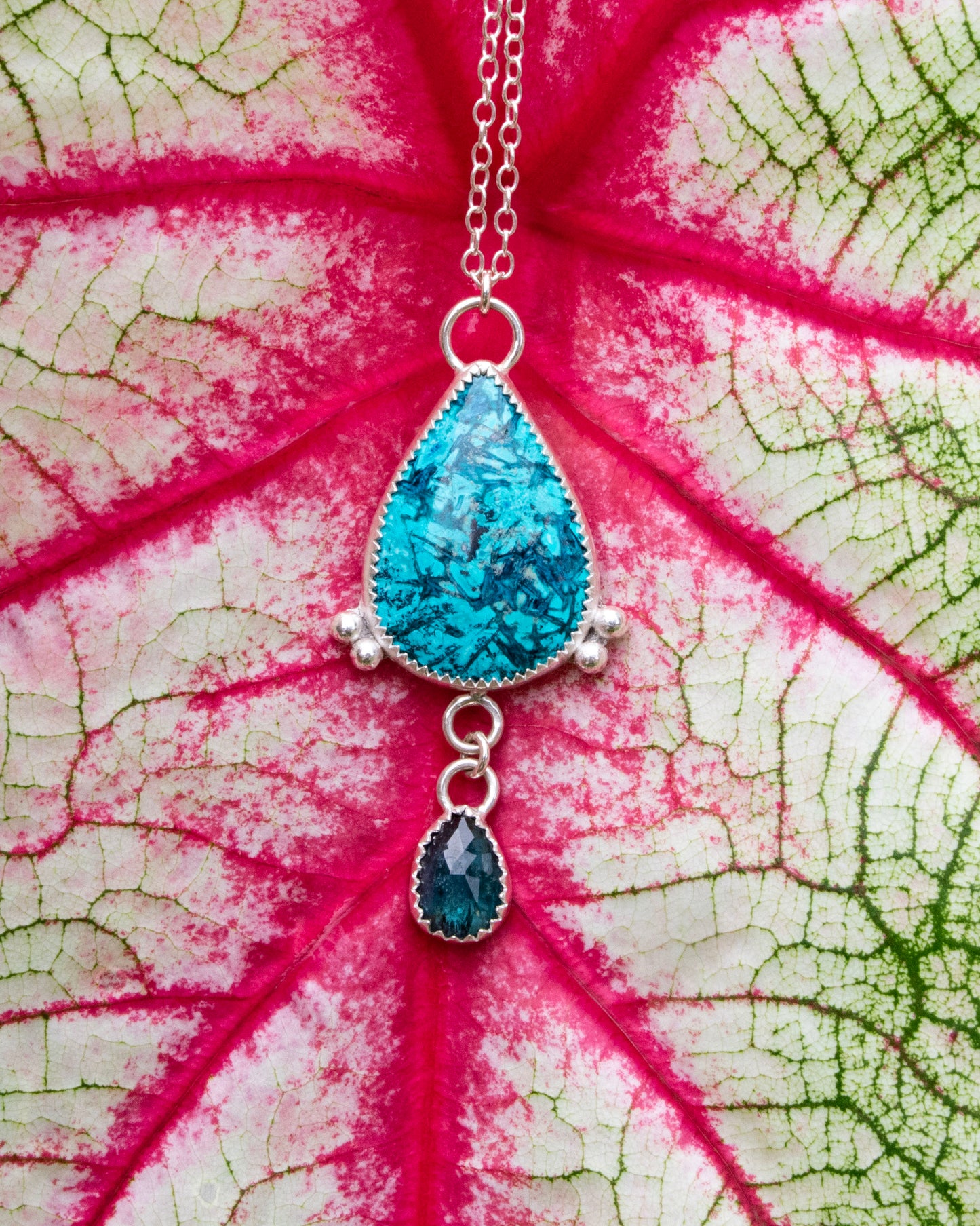sterling silver necklace with teardrop Chrysocolla with light and dark teal blue striation pattern with two silver beads at the bottom corneres of stone. 3 jumprings connect a small teal moss kyanite hangs from the bottom. Necklace is in front of red veins on white and green leaf