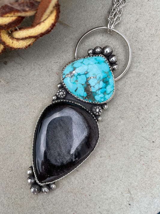 white water turquoise and silver sheen obsidian handcrafted necklace with silver accent beads hung from a large circle bail on silver french rope chain sitting on a felt hat background