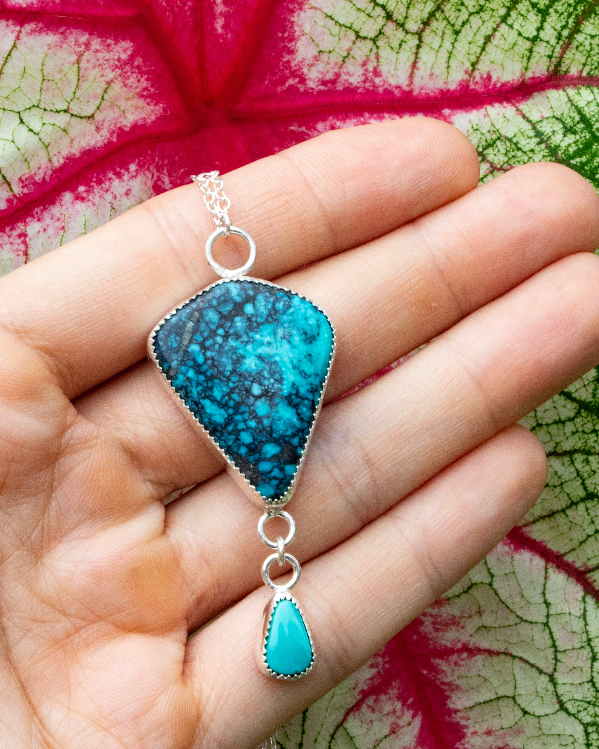 Moon River Turquoise kite sterling silver necklace with carico lake turquoise accent hung from a short jump ring chain at bottom of moon rivere stone. Necklace is being held in front of red veins of white and green leaf 