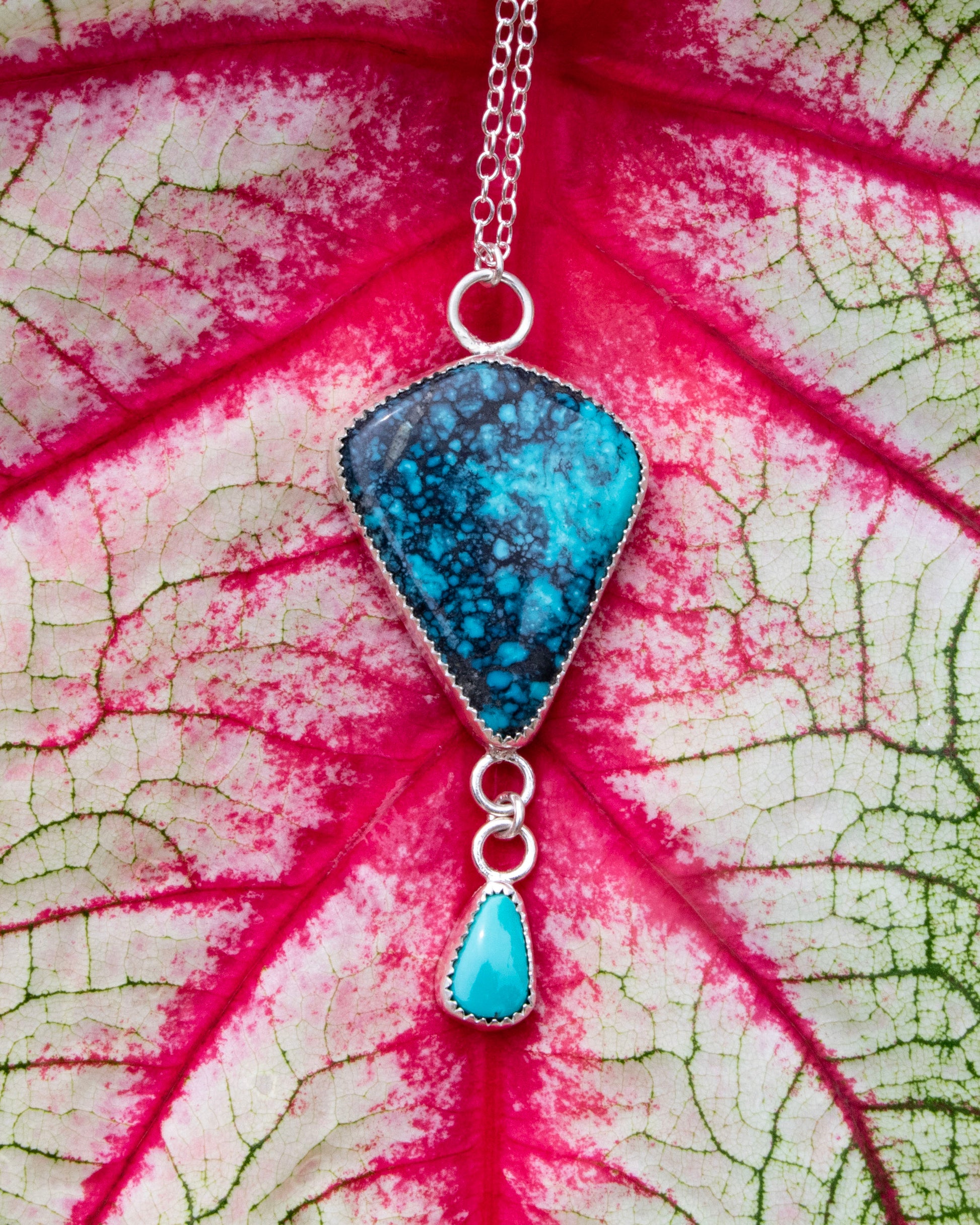 Moon River Turquoise kite sterling silver necklace with carico lake turquoise accent hung from a short jump ring chain at bottom of moon rivere stone. Necklace is sitting in front of red veins of white and green leaf 
