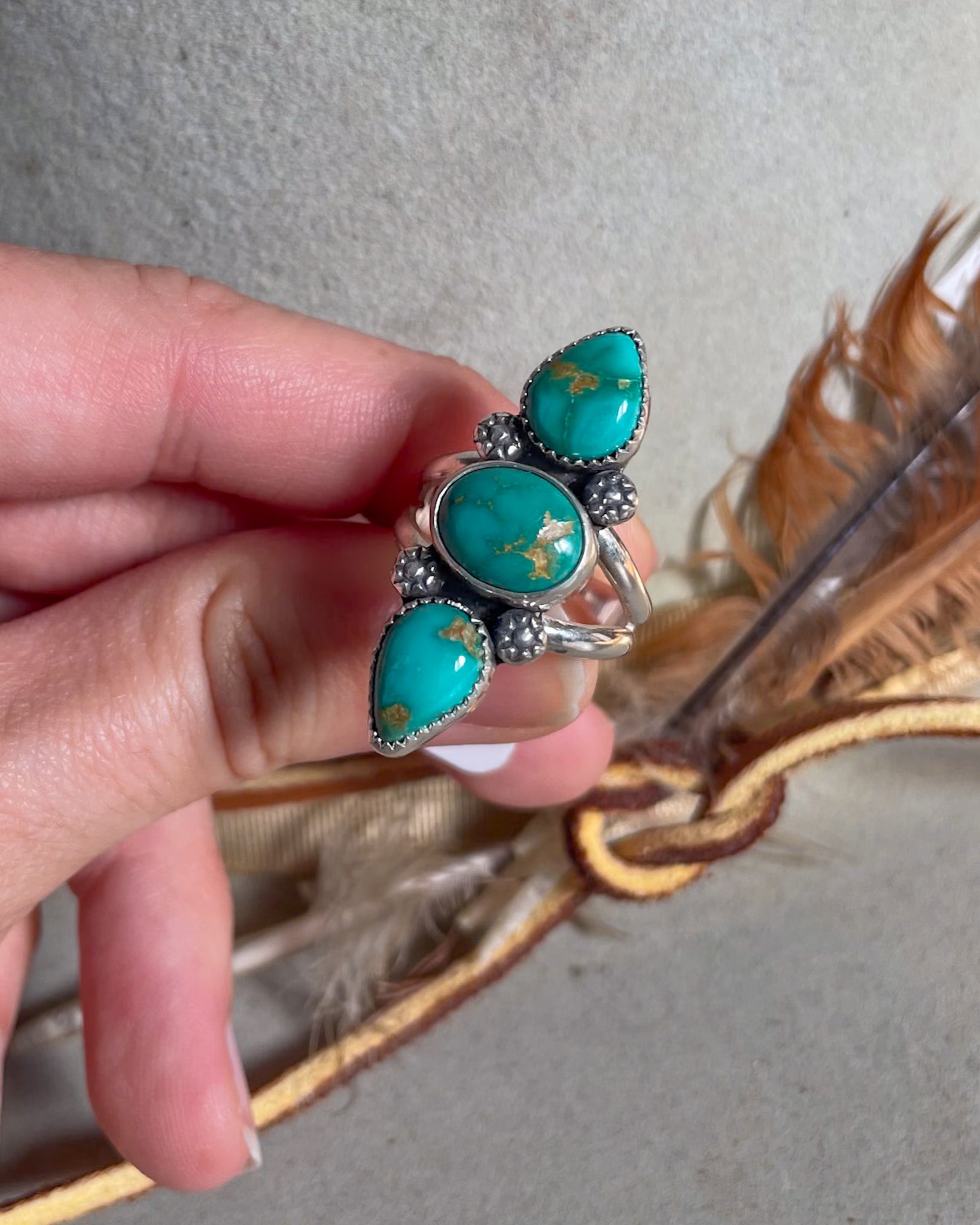 Turquoise ring with silver accents held by a hand, with feathers in the background